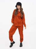 Woman in rust orange jumpsuit wearing black cloche hat, standing pose with hand raised